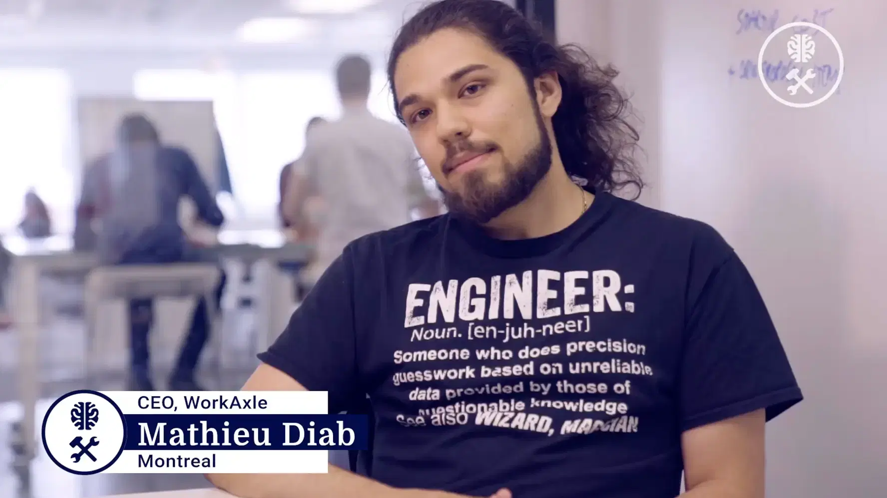 Mathieu Diab, CEO of WorkAxle in Montreal, sitting indoors with a relaxed posture, wearing a black T-shirt with humorous engineer definition text, with a blurred office background